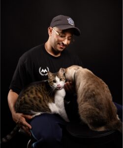 A smiling man in a baseball cap sits against a dark backdrop while holding two cats in his lap.
